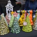 Ceramiczne choinki w zieleni z gwiazdkami stoją na czarnym stole. W tle kolorowe figurki i biała latarnia. Atmosfera targu.