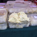 Plastikowe pudełka z parowanymi pierogami na zielonym tle. Pierogi w kolorze jasnokremowym. Skraplająca się para na pokrywkach.