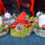 Świąteczne stroiki z czerwonymi i białymi świecami, szyszkami i gałązkami na ciemnym tle. Dominują kolory czerwony i zielony. W tle osoby i niebieskie butelki.