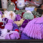 Na zdjęciu widać kolorowe ręcznie robione czapki z włóczki w odcieniach fioletu i różu. W tle są zielone ozdoby z wełny. Całość umieszczona jest na stoisku na jasnym tle.
