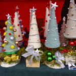 Na zdjęciu są ręcznie robione choinki z sznurka, ozdobione koralikami. Tło czerwone, jarmark bożonarodzeniowy. Detale: bombki, szyszki.