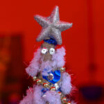 Dekoracyjna choinka z brokatową gwiazdą na górze, ozdobiona niebieskimi cekinami i złotymi kulkami. Tło w intensywnym czerwonym kolorze. Świąteczny akcent.