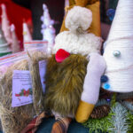 Na zdjęciu widać świąteczne ozdoby: kolorowe choinki z tkanin oraz paczki z sianem. Przeważają ciepłe i jasne barwy z dodatkiem zieleni. W tle dekoracyjne stożki i igliwie.