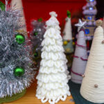 Na drewnianym stole stoją ozdobne choinki z jasnych materiałów. Tło jest czerwone. Niektóre choinki mają ozdoby, jak zielone kule i srebrne gwiazdy.