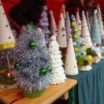 Stół z kolorowymi, ręcznie robionymi dekoracjami świątecznymi w kształcie choinek. Tło czerwone, atmosfera targowa. Detale ozdobione bombkami i wstążkami.