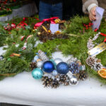 Stół z białym obrusem pełen zielonych wieńców świątecznych. Ozdoby z szyszek, kolorowych bombek i czerwonych wstążek. W tle widać ręce układające dekoracje.