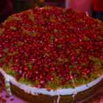 Ciasto pokryte zielonymi okruchami, przyozdobione czerwonymi ziarnami granatu. Tło w odcieniach różu. Całość wygląda apetycznie.