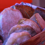 Kosz z różnorodnymi chlebami na czerwonym tle. Pieczywo ma złocistą skórkę, znajduje się w kolorowej ściereczce. Widoczny rustykalny charakter bochenków.