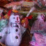 Na zdjęciu widoczne są ręcznie robione, kolorowe bałwanki w koszyku. Przykryte są przezroczystą folią z czerwonymi wstążkami. Tło to drewniana powierzchnia z różnymi opakowaniami.