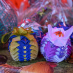 Na zdjęciu widać kolorowe, ręcznie robione ozdoby z materiału. Dominują barwy niebieska, złota, fioletowa i różowa. Tło jest rozmyte, z detalami sugerującymi lokalny targ lub stoisko.