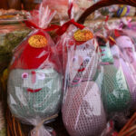 W wiklinowym koszu znajdują się kolorowe, zapakowane na szydełku maskotki. Mają różne kolory i nakładki z twarzami. Tło jest rozmyte, z pomarańczowymi i brązowymi odcieniami.