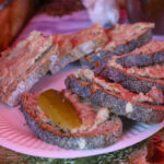 Kromki chleba z pastą i ogórkiem na białym talerzu. Dominują ciepłe kolory, w tle drewniane powierzchnie i wzory.