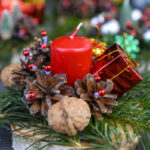 Czerwona świeca na tle zielonych gałązek świerku. Otoczona szyszkami i małym czerwonym prezentem. Tło rozmyte, świąteczne detale.