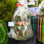 Ozdoby świąteczne w folii z czerwonymi i złotymi bombkami na tle zielonego obrusu w paski. Obok zielony wieniec. W tle choinka z lampkami.