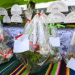 Na zdjęciu widoczne są zapakowane kosze z ozdobami świątecznymi, owinięte celofanem z czerwonymi i zielonymi wstążkami. W tle znajduje się stół z kolorowym obrusem i papierowymi talerzami z napisami. Dominuje kolor ziel