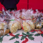 Na zdjęciu widać opakowane ciasteczka i orzechy w plastikowych woreczkach z kolorowymi wstążkami. Tłem jest czerwone materiałowe stoisko, a stół przykryty jest bożonarodzeniowym obrusem w ostrokrzewy. Na stole leży również