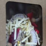 Pudełko z przezroczystym okienkiem zawiera ozdoby świąteczne. Widać kremowe wstążki i czerwone akcenty. Tło kartonowe.