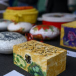 Zdjęcie przedstawia kolorowe, ozdobne pudełka i jajka na ciemnym stole. Dominuje żółć, zielenie, czerwienie i fiolety. Tło jest rozmyte.