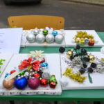 Na zielonym stole leżą kolorowe dekoracje świąteczne z bombkami i girlandami. Są też kartki z tekstem i krzesła w tle. Dominują srebrne, złote i czerwone ozdoby.