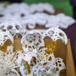 Na zdjęciu widać białe, szydełkowe parasolki w kartonie. Tło jest rozmyte z zielonymi i fioletowymi akcentami. Parasolki ozdobione są srebrnymi i złotymi elementami.