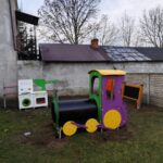 IMG_20251205_133308 Kolorowy plac zabaw z lokomotywą stoi na trawie. W tle ściana budynku i płot z cegieł. Obok znajduje się tablica i kuchnia zabawkowa.
