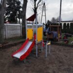 IMG_20251205_133322 Plac zabaw z czerwoną zjeżdżalnią i żółtym panelem na tle drzew i płotu. Ziemia dookoła jest nieprzykryta trawą. Obok placu leży sprzęt ogrodowy.
