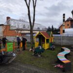 IMG_20251205_133333 Plac zabaw z kolorową altanką, tęczową ławką i sprzętem do ćwiczeń. W tle osoby pracują i budynek z cegieł. Niebo pochmurne.