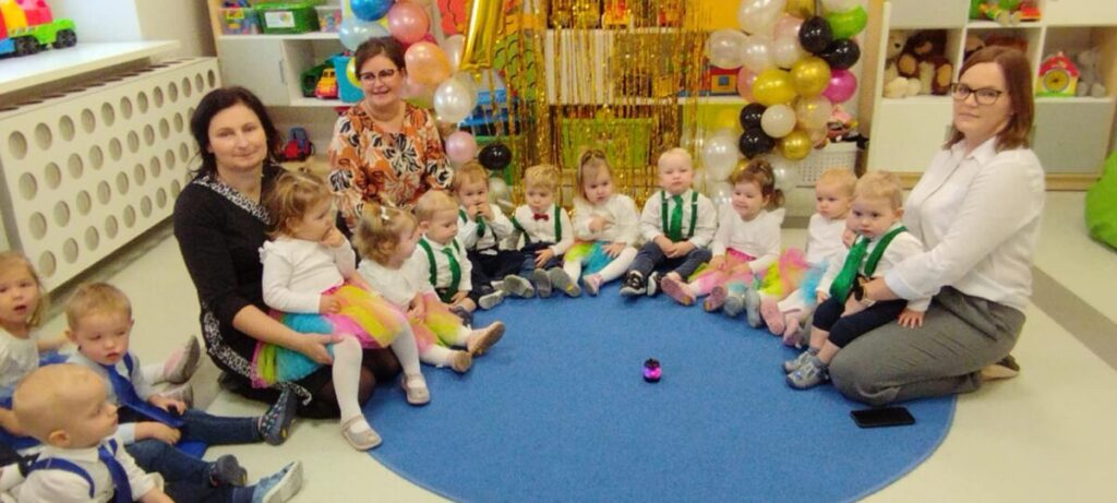 Grupa dzieci i opiekunów siedzi w kole na niebieskim dywanie. W tle kolorowe balony i zabawki. Wszyscy uśmiechnięci.