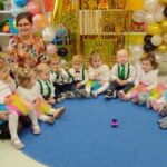 Grupa dzieci i opiekunów siedzi w kole na niebieskim dywanie. W tle kolorowe balony i zabawki. Wszyscy uśmiechnięci.