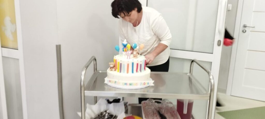 Kobieta w białej bluzce ustawiająca kolorowy tort na wózku. Tło jest jasne z białymi drzwiami. Tort ma dekoracje z balonami i liczbą 7.