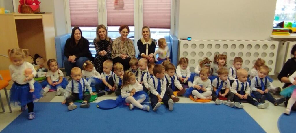 Grupa małych dzieci i cztery kobiety siedzą na dywanie. Dzieci mają eleganckie stroje, dziewczynki w niebieskich spódniczkach. Tło jest jasne, z zabawkami po lewej stronie.