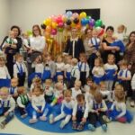 Na zdjęciu widoczna jest grupa dzieci i dorosłych w sali, ubrani w kolory niebieski i biały. Tło zdobi kolorowa girlanda z balonami. Widać również zabawki i dekoracje na ścianach.