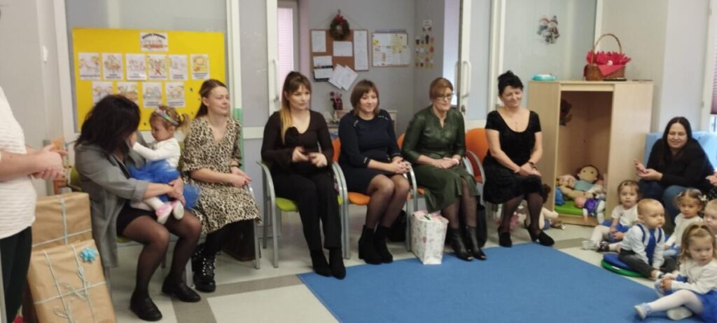 Na zdjęciu grupa dorosłych i dzieci w jasnym pokoju. Kobiety siedzą na krzesłach, dzieci bawią się na niebieskim dywanie. W tle tablica z rysunkami i zabawki.