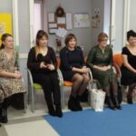 Na zdjęciu grupa dorosłych i dzieci w jasnym pokoju. Kobiety siedzą na krzesłach, dzieci bawią się na niebieskim dywanie. W tle tablica z rysunkami i zabawki.