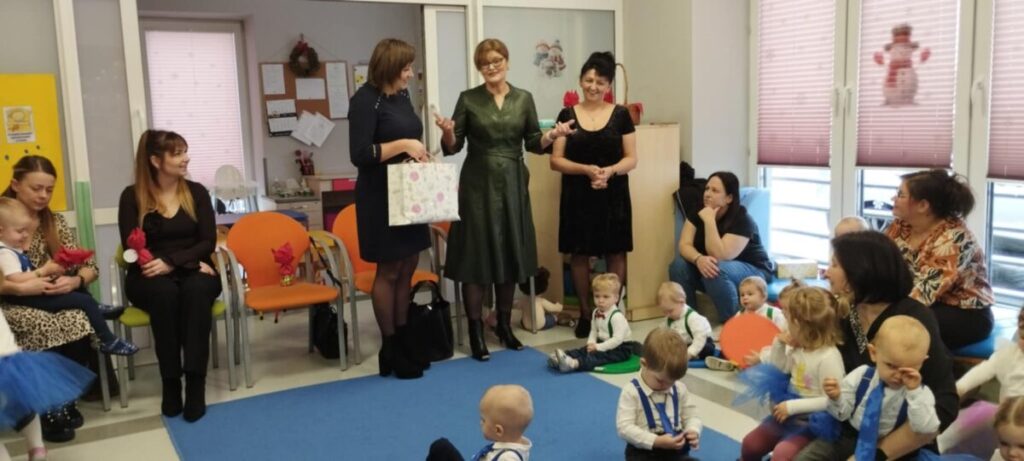 Grupa dorosłych i dzieci w kolorowej sali przedszkolnej. Na podłodze niebieska mata, a w tle krzesła i tablica. Dorośli stoją i siedzą, trzymając dzieci.