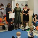 Grupa dorosłych i dzieci w kolorowej sali przedszkolnej. Na podłodze niebieska mata, a w tle krzesła i tablica. Dorośli stoją i siedzą, trzymając dzieci.