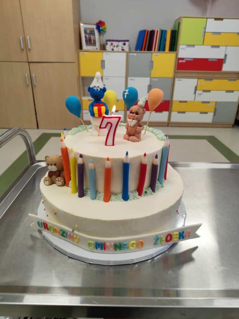 Tort z figurkami niebieskiego i beżowego stworka, kolorowe świeczki, cyfra 7 na świeczce. Tło: kolorowe szafki.