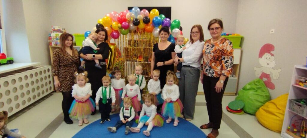 Grupa dzieci w kolorowych strojach z opiekunami stoi na niebieskiej macie. W tle balony i szare ściany. Obok zabawki i pluszowy Gargamel.