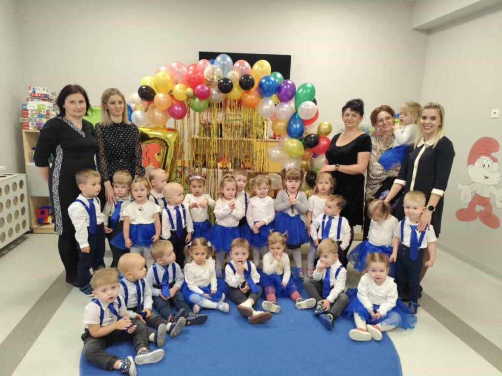 Grupa dzieci i nauczycielki w kolorowych strojach na tle balonów w sali. Dzieci mają niebieskie elementy ubrań. Pomieszczenie zdobi postać smerfa.