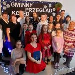 Na zdjęciu grupa osób na tle czarnego banera z napisem Gminny Przegląd. Ubrania w jasnych kolorach, w tym czerwona sukienka. Tło ozdobione światełkami i śnieżynkami.