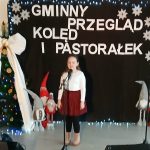 Dziewczynka śpiewa na czarnym tle z napisem Gminny Przegląd Kolęd i Pastorałek. Obok choinka i dekoracje świąteczne. Scena oświetlona lampkami.