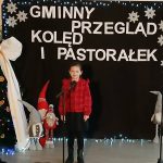 Dziewczynka w czerwonej bluzce stoi na scenie. W tle czarne płótno z białymi napisami „Gminny Przegląd Kolęd i Pastorałek”. Obok dekoracje świąteczne: choinka z białymi kokardami i figurki Mikołaj