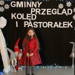 Dziewczynka w czerwonej sukience śpiewa na scenie. W tle czarny baner z napisem Gminny Przegląd Kolęd i Pastorałek. Obok ozdobiona choinka i figurki elfów.