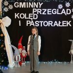 Dziecko śpiewa na scenie przed czarnym tłem z napisem Gminny Przegląd Kolęd i Pastorałek. Obok ozdobiona choinka i krasnale. Dekoracje świąteczne w bieli, zieleni i czerwieni.