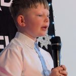 Chłopiec w białej koszuli trzyma mikrofon. Tło ozdobione białymi śnieżynkami i czerwonymi gałązkami. Dekoracje sugerują zimowy nastrój.