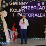 Chłopiec w białej koszuli śpiewa na tle ciemnego baneru z napisem Gminny Przegląd Kolęd i Pastorałek. Po lewej stronie jest ozdobiona choinka z białą kokardą, obok stoją figurki krasnali. T