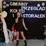 Kobieta śpiewa na scenie przy choince, z napisem „Gminny Przegląd Kolęd i Pastorałek” na czarnym tle. Dekoracje to śnieżynki i krasnale. Ubrana w czarną sukienkę i buty.