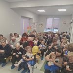 Na zdjęciu widać grupę dorosłych i dzieci siedzących w jasnym pomieszczeniu. Dorośli trzymają dzieci na kolanach, większość z nich je deser. W tle są rysunki na ścianie i dwa małe okna.