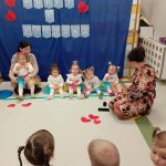 Dzieci w kolorowych strojach siedzą przed niebieskim tłem z napisem Dzień Babci i Dziadka. Na podłodze czerwone serduszka. Dwóch dorosłych towarzyszy dzieciom.