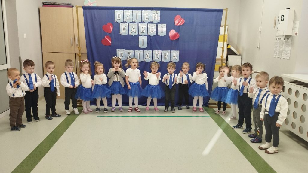 Grupa dzieci w białych koszulach i niebieskich spódniczkach stoi przed niebieską kurtyną. Napis Dzień Babci i Dziadka, czerwone serca. Dzieci są w sali z jasnymi ścianami i podłogą.